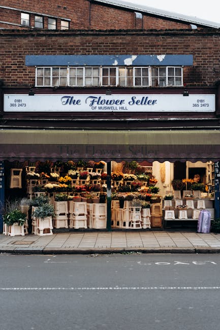 The exterior of The Flower Seller shop on Muswell Hill features a prominent white sign with elegant blue lettering, indicating its specialization in flowers. The shopfront displays a vibrant arrangeme