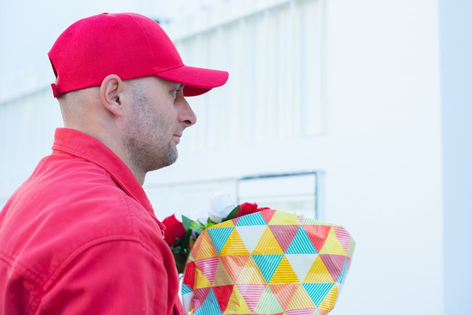 A man wearing a red cap and red jacket is holding a large, colorful gift box wrapped in paper with a geometric triangle pattern in shades of yellow, blue, red, and green. The background features a lig