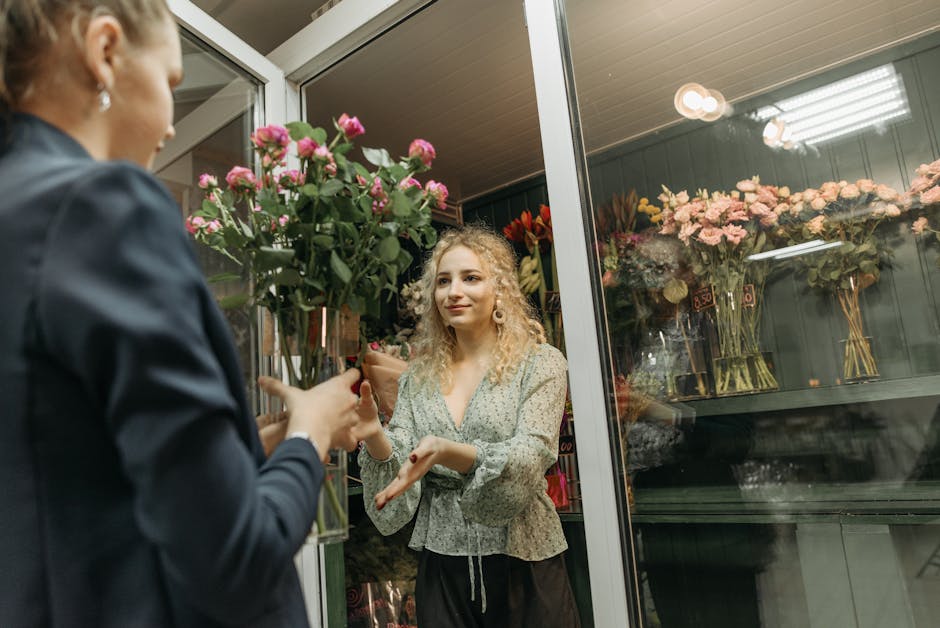 A young woman with curly blonde hair, dressed in a light floral blouse, is inside a flower shop near New Malden Station, receiving a bouquet of fresh pink roses wrapped in brown paper from a florist. 