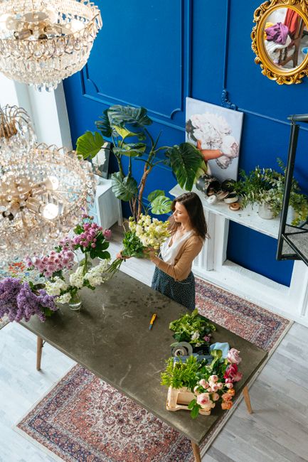 A woman arranging a fresh bouquet of white and yellow flowers on a large, dark grey table inside a vibrant blue-walled floral studio. The scene features an ornate chandelier hanging from the ceiling, 