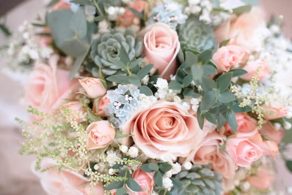 A close-up view of a floral arrangement featuring a mix of soft pink roses, small pink spray roses, and pale blue succulents, all surrounded by green eucalyptus leaves and delicate white baby's breath