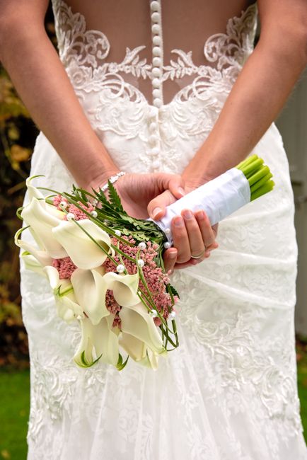 Close-up of a bride holding a bouquet of fresh white calla lilies, pink astilbe, and green foliage, wrapped in white ribbon, with her hands gently clasped around the stems. She wears a lace wedding go