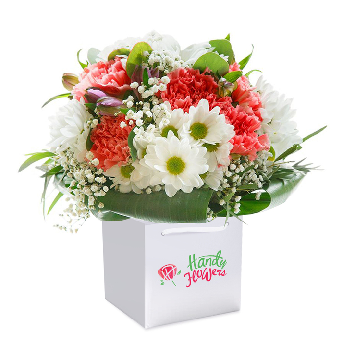 This beautifully arranged bouquet, upright within a crisp square white gift bag, radiates with the colour and vibrancy that New Malden residents cherish in local markets and home gardens. Ruffled coral, peach, and red carnations take centre stage, their soft, full petals conveying joy and warmth, complemented gracefully by snowy white daisies sporting zesty green centers. Each flower is thoughtfully layered, interspersed with delicate sprays of baby's breath-tiny white blooms that evoke a sense of lightness and charm. Broad, deep green glossy leaves shape a lush base, embracing the blooms with a flourish that mirrors the abundant lawns of Beverley Park. Tucked discreetly at the side, unopened purple-tipped buds promise new beginnings and lend a sophisticated edge to the display. The bag's sturdy ribbon handles and subtle curves suggest care, attention, and functionality, perfect for safe and elegant flower delivery around New Malden's High Street. Bathed in flattering natural light, the entire arrangement appears fresh and exuberant, designed to mark birthdays, anniversaries, or heartfelt gestures with a truly local touch. In every detail, from the curated colour palette to the softness of the foliage, this gift embodies the thoughtful craftsmanship that defines New Malden's best florists.
