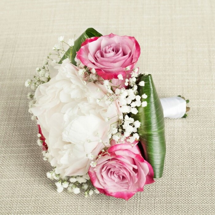 From the creative hands of New Malden's seasoned florists comes this compact yet lavish bouquet-ideal for a bridesmaid or as a centerpiece for life's most meaningful celebrations on Kingston Road and beyond. The bouquet features two luminous pink roses, each petal delicately spiraled and brushed with a gradient from vivid magenta at the outer edge to soft, romantic blush and hints of lavender. Their movement guides the eye toward the central white peony, whose densely layered, ruffled petals impart a feeling of opulence and gentle luminosity under natural, even lighting. Sprigs of gossamer baby's breath scatter throughout the bouquet, introducing a celestial, almost dreamlike dimension that captures both light and wonder. Encircling these blooms, broad, rich green leaves shape a structural base, folded expertly and finished with a crisp white ribbon that forms a graceful handle. The bouquet, presented on a textured beige linen reminiscent of New Malden's welcoming homes and venues, exudes an air of thoughtful elegance and joy-perfect for marking new beginnings or celebrating community bonds in this lively London suburb.