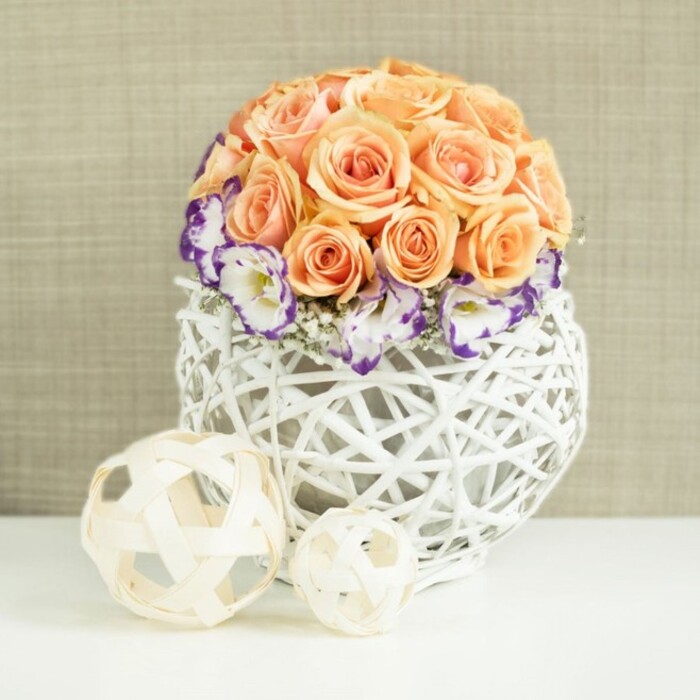 This image shows a refined and romantic floral centerpiece arranged with the care and precision you might expect from an experienced New Malden florist preparing a wedding table. The focal point is a lush dome of peach and apricot roses, closely clustered into a perfect sphere so that the viewer sees almost nothing but petal. Each rose appears fully in bloom, the velvety petals swirling outwards in tidy spirals, with tones that move gracefully from delicate, pale peach near the edges to richer apricot shades nearer the center, giving the bouquet depth and warmth. At the base of this rose dome, a graceful collar of white lisianthus flowers flares gently outward. Their soft, ruffled petals are edged with deep violet and purple, creating a crisp line of colour that defines the bottom of the rose globe and adds a sophisticated contrast. Seated between the lisianthus stems, fine clusters of white gypsophila-baby's breath-create tiny bursts of softness, lending a light, cloud-like effect around the base of the arrangement. The flowers are gathered into a distinctive, spherical white vase woven from slender rattan or wicker strands, forming an open lattice that allows the stems and inner shadows to become part of the design. In front of the vase, on the same reflective white surface, sit two decorative balls in pale, natural wood tones, each one a loosely woven sphere that mirrors the curvature and texture of the container while adding a sculptural touch. Behind it all, a softly textured beige backdrop, reminiscent of linen or burlap, provides a neutral canvas that lets the warm peach and cool purple tones shine. The mood is tranquil and celebratory, perfect for an intimate wedding in New Malden, a special anniversary dinner at home, or a carefully styled photo shoot showcasing elegant, contemporary floral design.