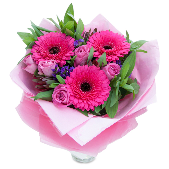 The charm of this pink bouquet lies in its contrast of playful energy and quiet grace: three hot-fuchsia Gerbera daisies burst forward with layered petals circling deep dark centres, while soft lavender-pink roses, still snug in early bloom, provide a soothing, romantic touch. Tiny sprays of purple statice weave through the larger flowers like flecks of evening sky, offering a delicate counterpoint and pleasing tactile variety. Surrounding the blooms are broad, glossy green leaves - likely salal or eucalyptus - chosen both for visual fullness and their clean, slightly dewy scent that mingles with the roses' subtle perfume. The flowers rest on a base of soft pale pink tissue paper and are wrapped in a sheer, slightly crinkled fuchsia-pink cellophane that adds a light, shimmering veil; the wrapper is folded with artisan care so the bouquet sits above the visible rim of a clear glass vase, hinting that it's ready to be placed on a mantle or table. Bathed in soft, neutral light against a white backdrop, the arrangement feels intimate and uplifting, an ideal present for a birthday, anniversary or to brighten a friend's day. As a florist familiar with New Malden - from Kingston Road shoppers to commuters arriving at the station - I picture this bouquet gracing kitchen tables and bedside stands across the neighbourhood, bringing the garden's sweetness indoors.