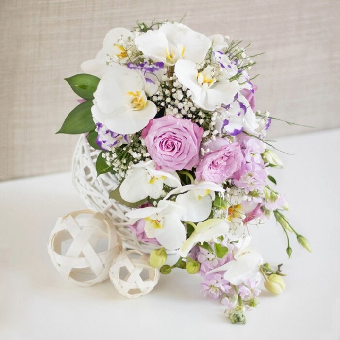A beautifully balanced floral centerpiece, crafted with the care of an experienced New Malden florist, rests on a spotless white surface against a softly blurred beige backdrop. The main focus is a sculptural white globe vase, created from interlaced strands that form an open, woven lattice reminiscent of delicate wicker work. From this airy base emerges a lush bouquet dominated by white Phalaenopsis orchids, their broad petals arching outward with quiet grace. Each orchid has a luminous quality, and at its centre a touch of yellow and pale amber adds gentle warmth, suggesting both purity and sophistication. Encircling the orchids are roses in muted blush pink and cool lavender tones, their petals layered and slightly ruffled, many blooms captured at the moment of perfect openness that evokes romance and celebration along the streets of New Malden. Between these feature flowers, smaller white petals tipped with purple or fuchsia-likely Lisianthus-create subtle shadows and highlights, bringing depth to the design. Fine sprays of gypsophila drift through the bouquet like a soft veil, adding volume without heaviness, while slender stems of light lilac flowers trail over the rim, introducing a natural cascade. Pockets of glossy green foliage are dotted throughout, framing the pastel tones and grounding the arrangement with fresh, garden-like energy. On the table beside the vase, two decorative cream spheres, woven in the same open style, echo the vase's lattice and complete the harmonious composition. The lighting is bright and even, revealing every texture from the orchids' satin-smooth petals to the roses' velvety folds, and together with the neutral background, it heightens the sense of calm luxury ideal for celebrations across New Malden.