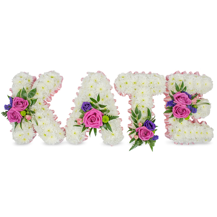 This elegant floral name tribute spells 'KATE' in bold, freestanding letters, each one fashioned from a compact sea of immaculate white chrysanthemums that form a smooth, cushion-like surface. A pale pink satin ribbon trims every edge, lending a refined, almost luminous outline to the capital letters against a stark white studio background. Rich fuchsia-pink roses form the focal points of several small clusters, accompanied by delicate pastel pink spray roses and touches of deep purple or blue filler blooms - likely statice or small lisianthus - which together create a measured, comforting palette of colour. Sprays of fern and eucalyptus tuck around each cluster, their varied textures lending dimension and a soft, aromatic freshness. The bloom placement reads like a quiet conversation: the diagonal arm of the 'K' holds a dense cluster, the 'A' receives flowers near its lower right curve, the 'T' is anchored with a bouquet at its base, and the 'E' is graced with two distinct groupings on its middle bar and bottom-each chosen to balance form and sentiment. The styling is restrained and reverent, suited to a funeral service, graveside tribute, or a celebration of life where family prefer a personalised and dignified expression. As a local floral piece, it would sit gracefully at a service near Kingston Road or on New Malden High Street, reflecting the careful craftsmanship and sensitivity of an experienced florist.